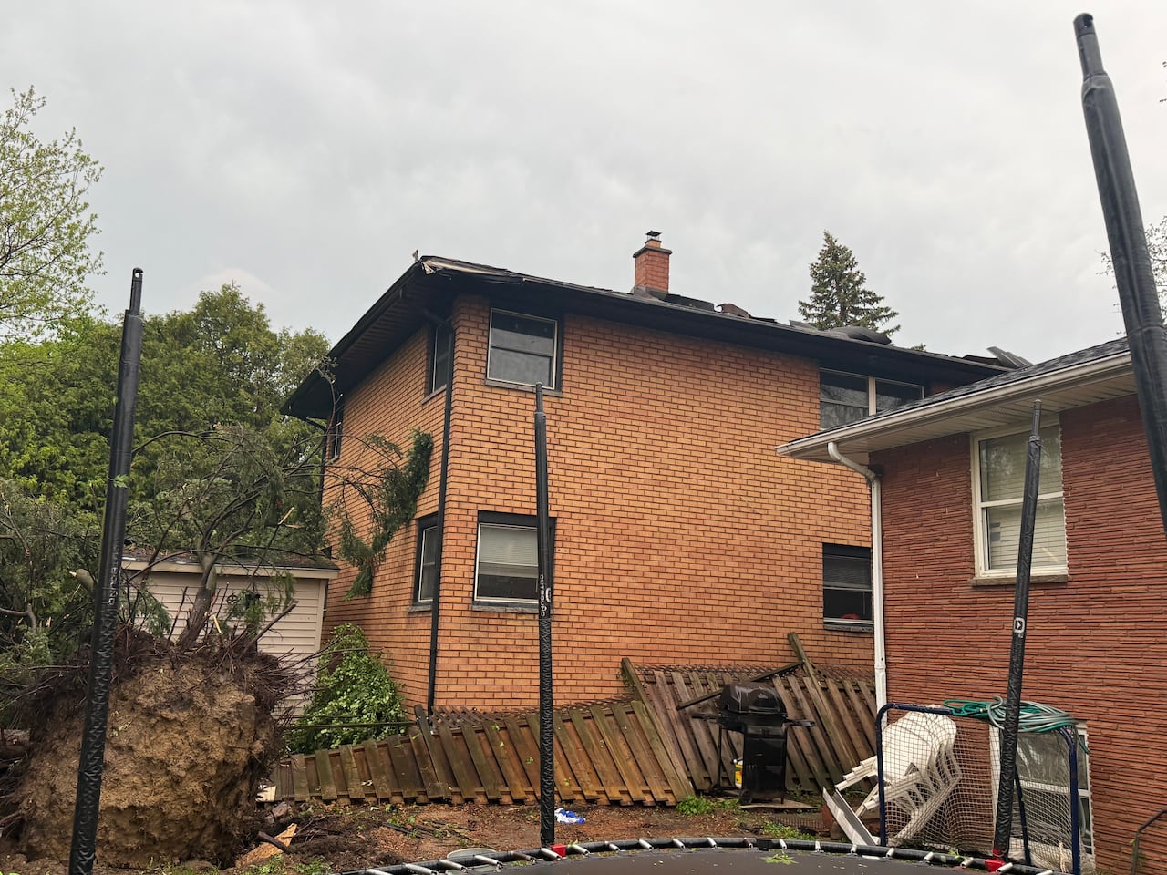 A house with a large uprooted tree in the yard, as well as a knocked over fence and roof damage