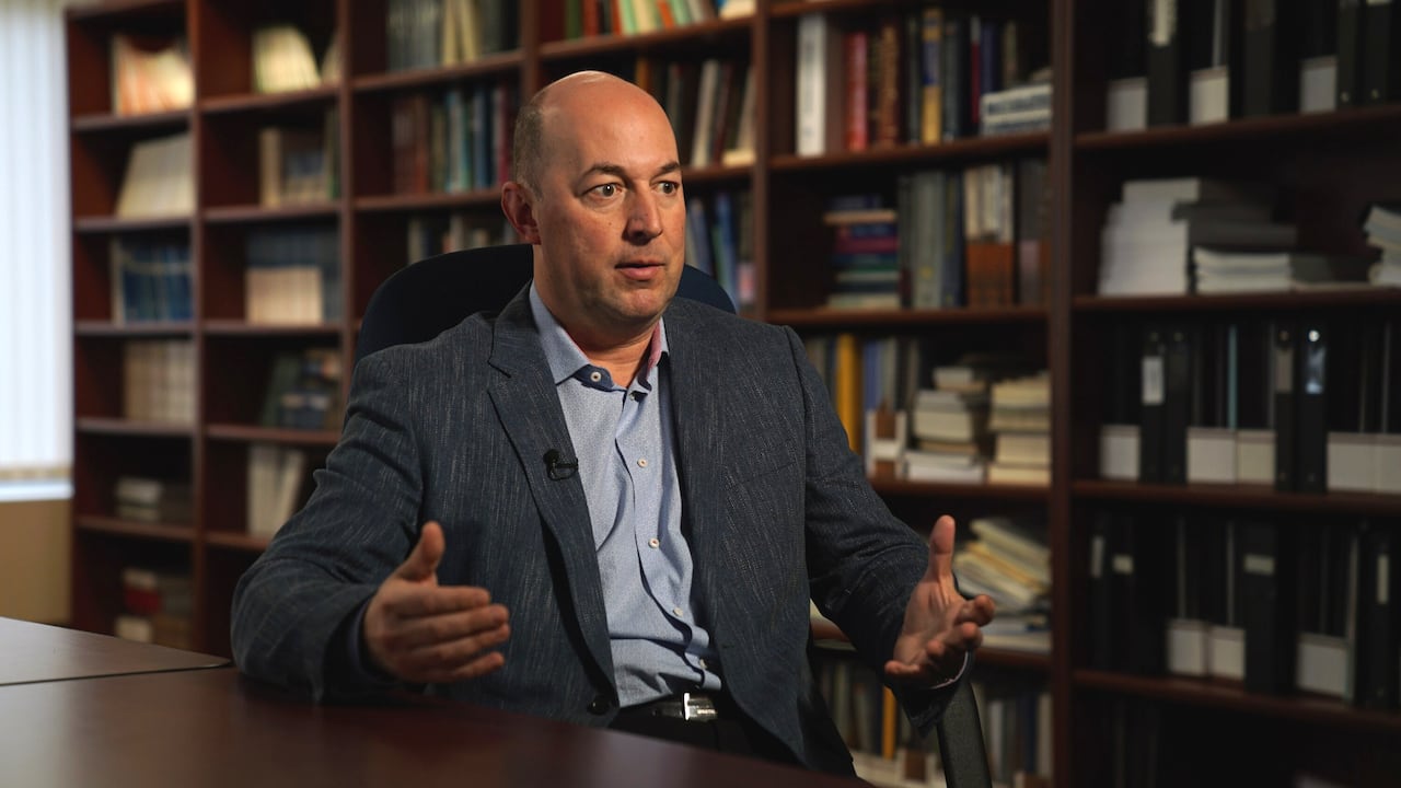 A portrait of a man in a dark blue suit jacket behind a bookcase.