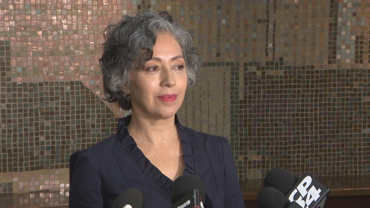 Woman in blue shirt stands in front of microphones smiling