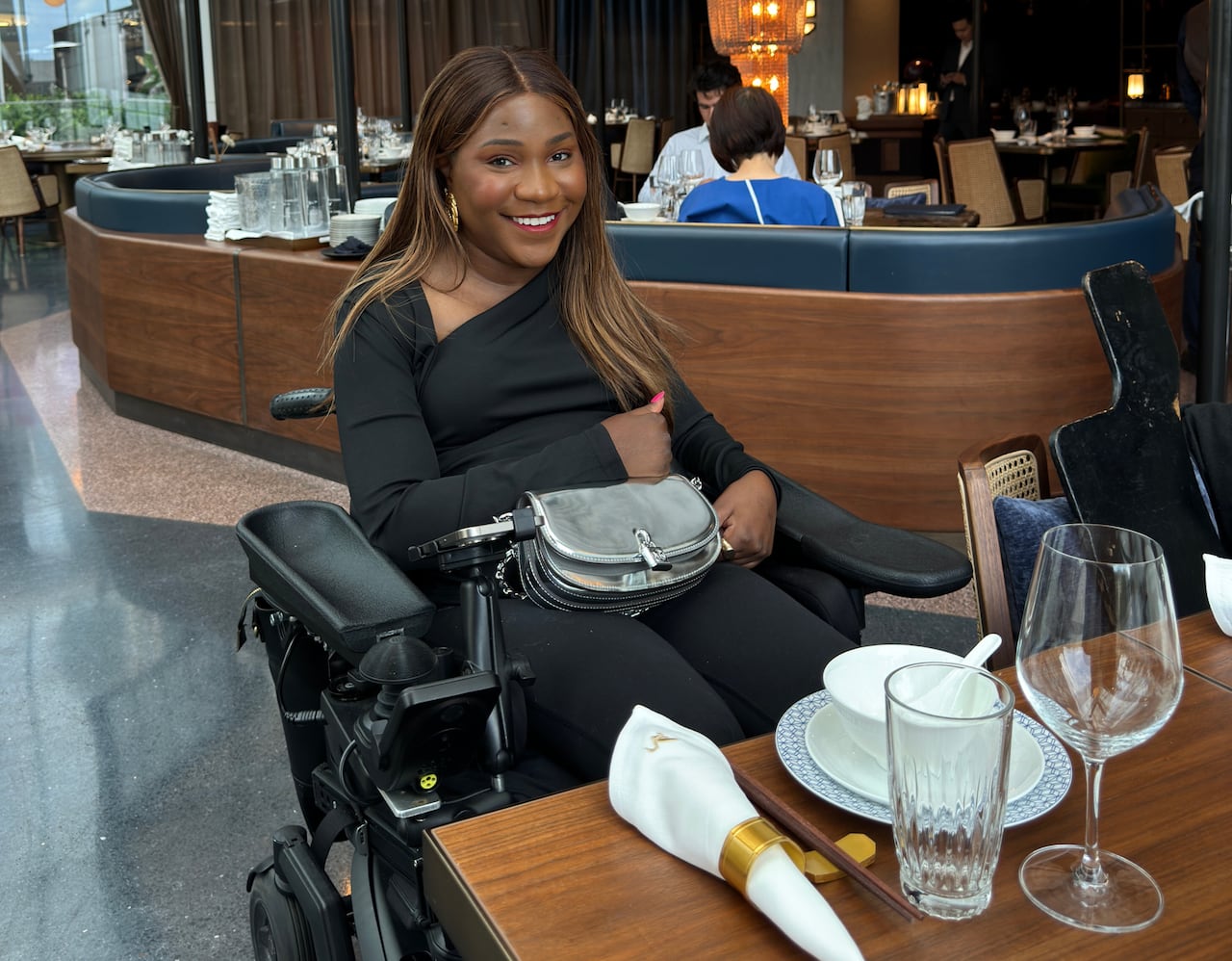 A woman in a wheelchair wearing all black smiles, sitting in front of a restaurant table with a silver purse on her lap.