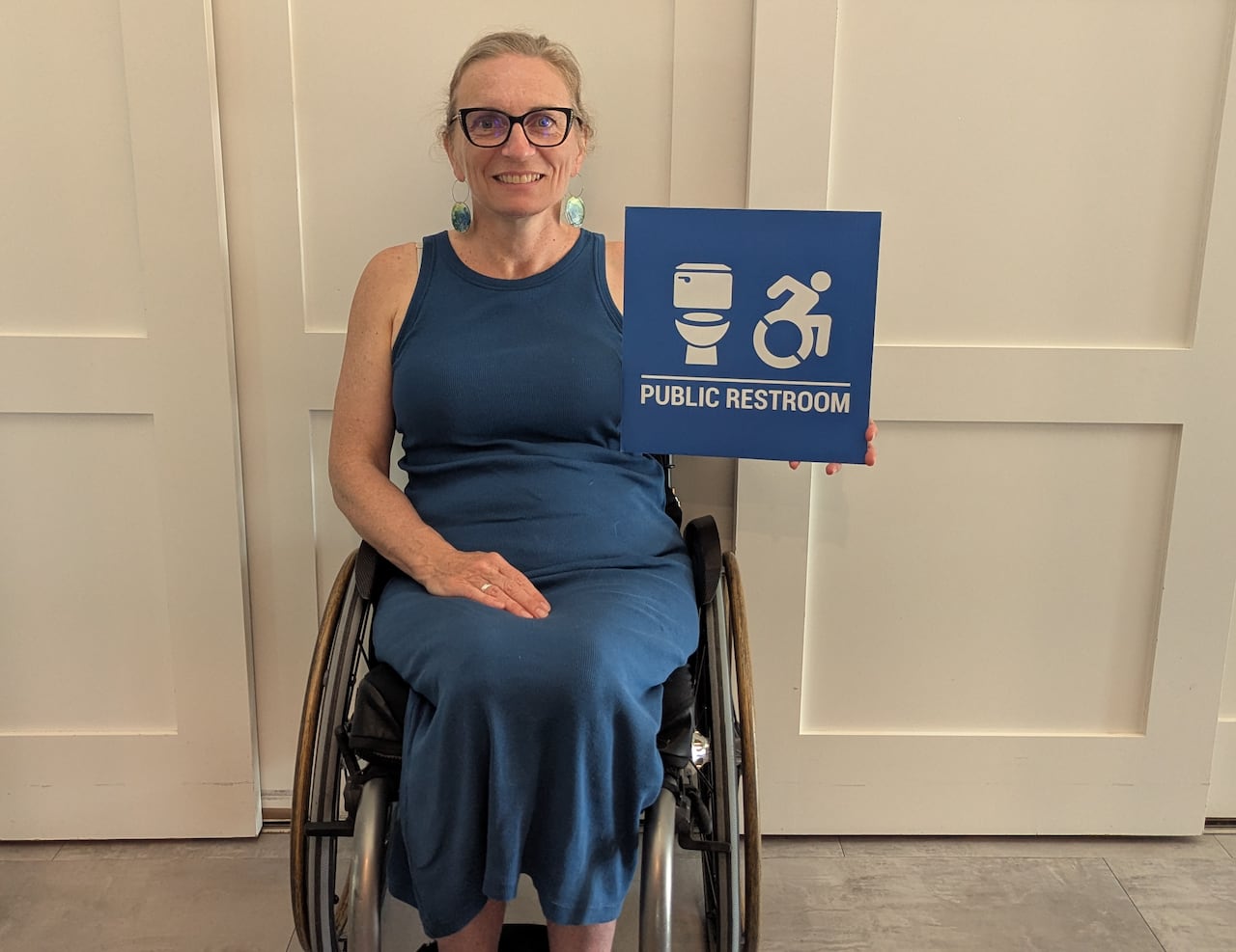 A woman in a wheelchair wearing a blue dress holds an accessible washroom blade sign.