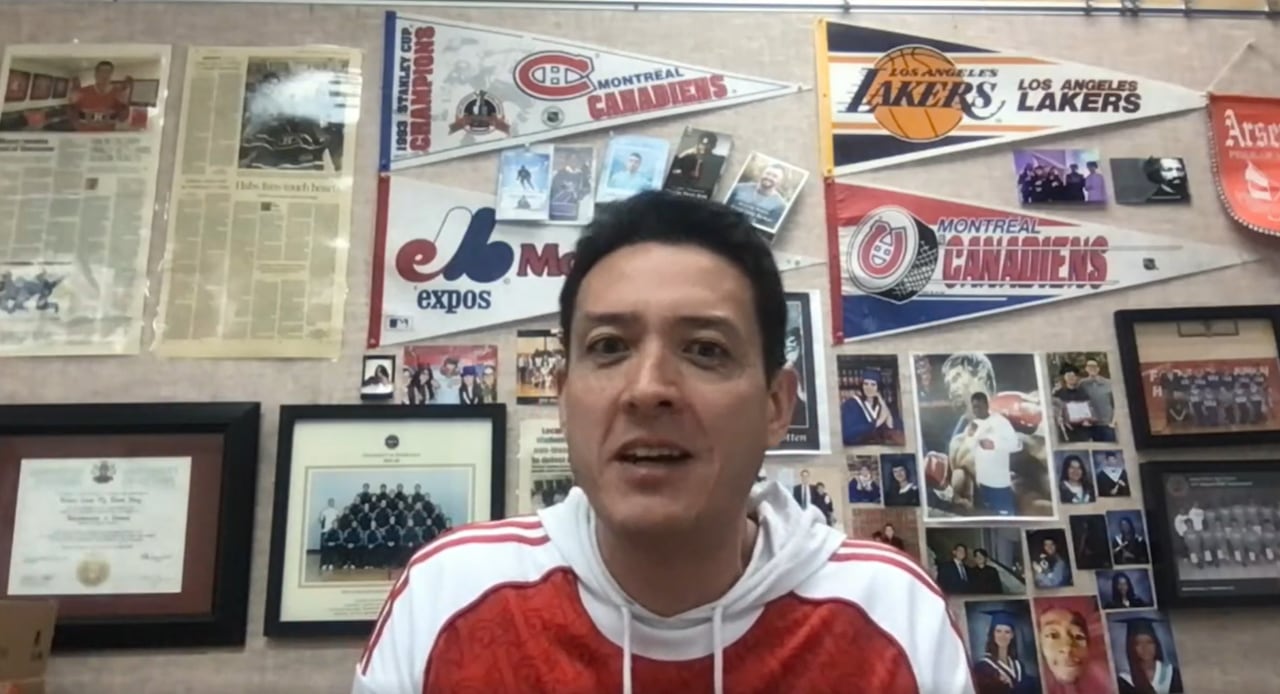 A man in a sports hoodie speaks on camera with a background consisting of sports memorabilia.