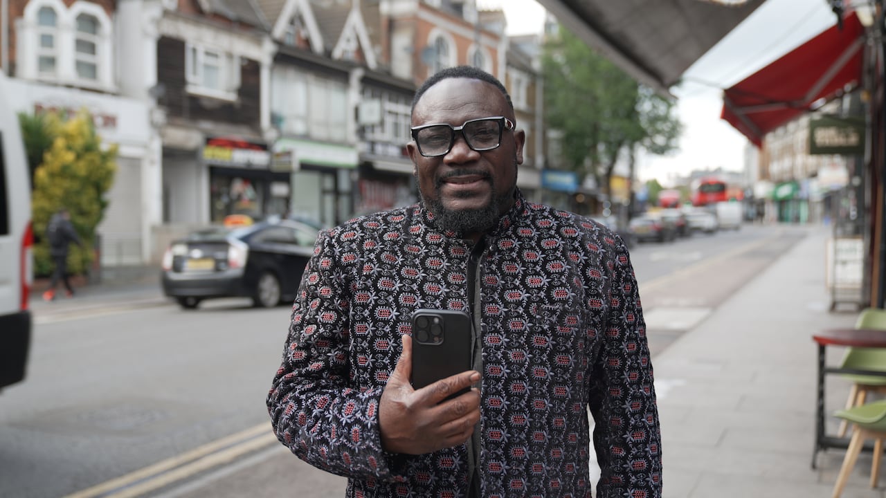 A man in a colourful jacket stands on a sidewalk in a neighbourhood in north London holding a new iphone.