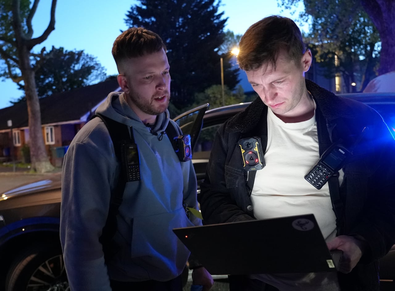 Two men with short hair wear radios and cameras and look at a laptop while standing outside a car with its door open.
