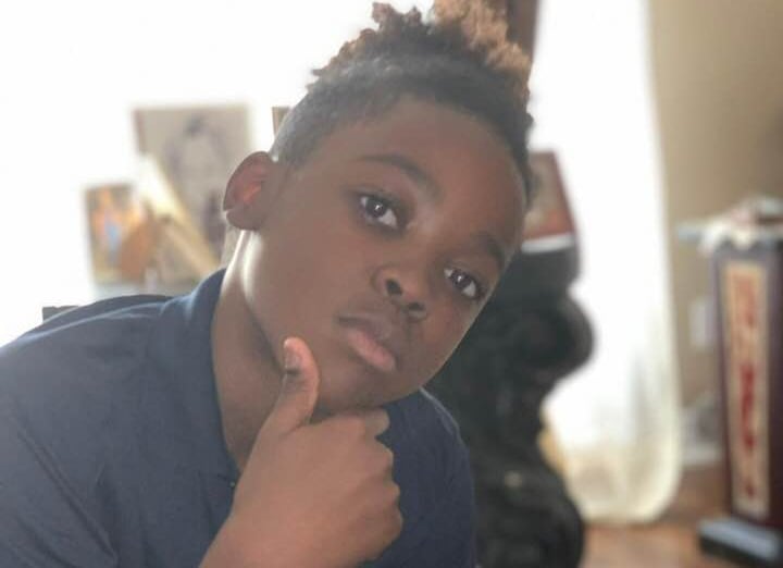 A young Black boy wearing a navy shirt holds his face as he looks seriously at the camera. 