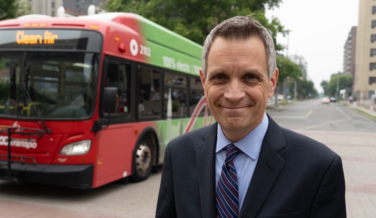 A man in a suit and tie stands in front of a bus and smiles.