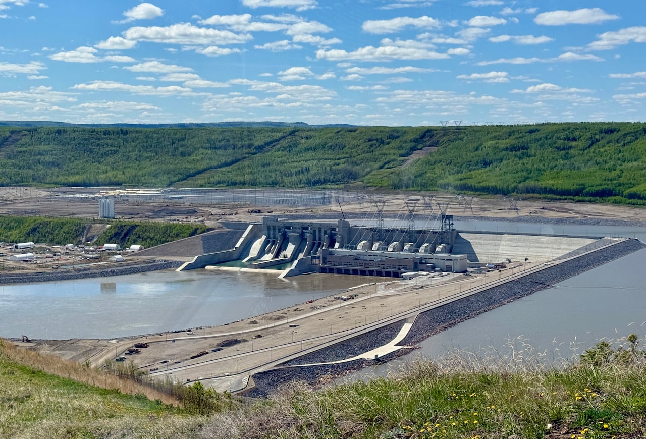 A giant hydroelectric dam spans a wide river, surrounded by lush green hills under a bright blue sky with scattered clouds.
