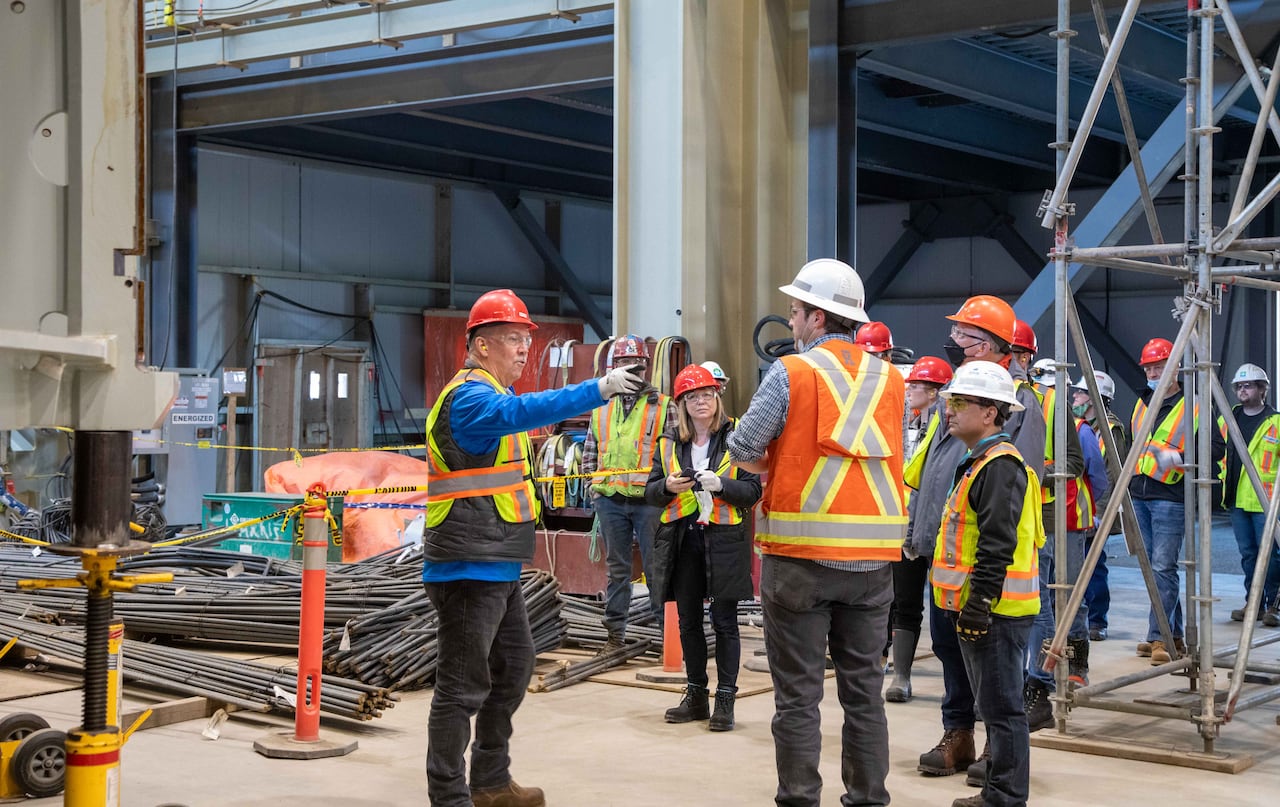 A group of construction workers wearing hard hats and reflective vests gathers inside a large industrial site as one person gestures and points.