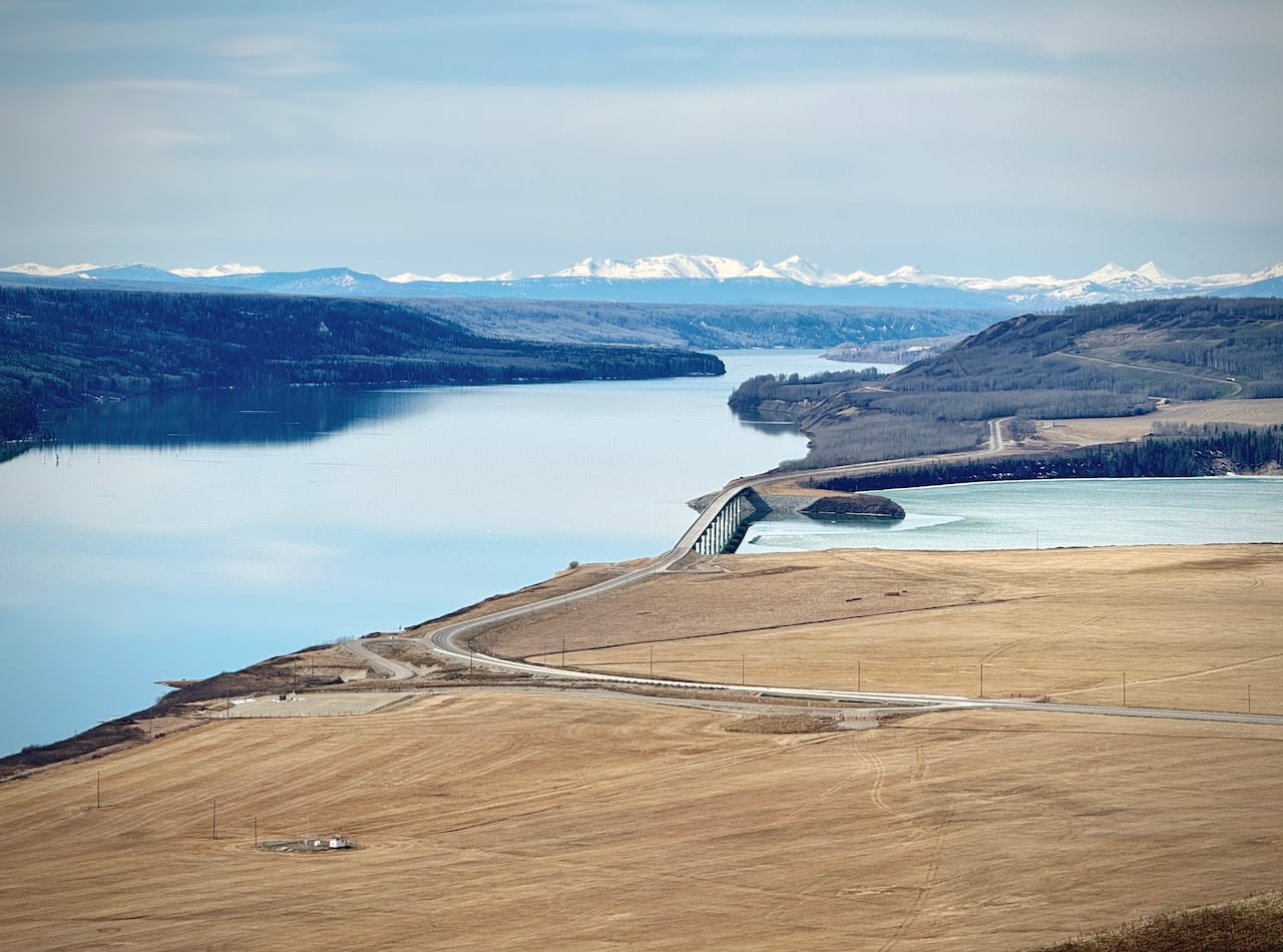 A landscape of a calm lake reservoir next to farm fields with a bridge and mountains in the background.
