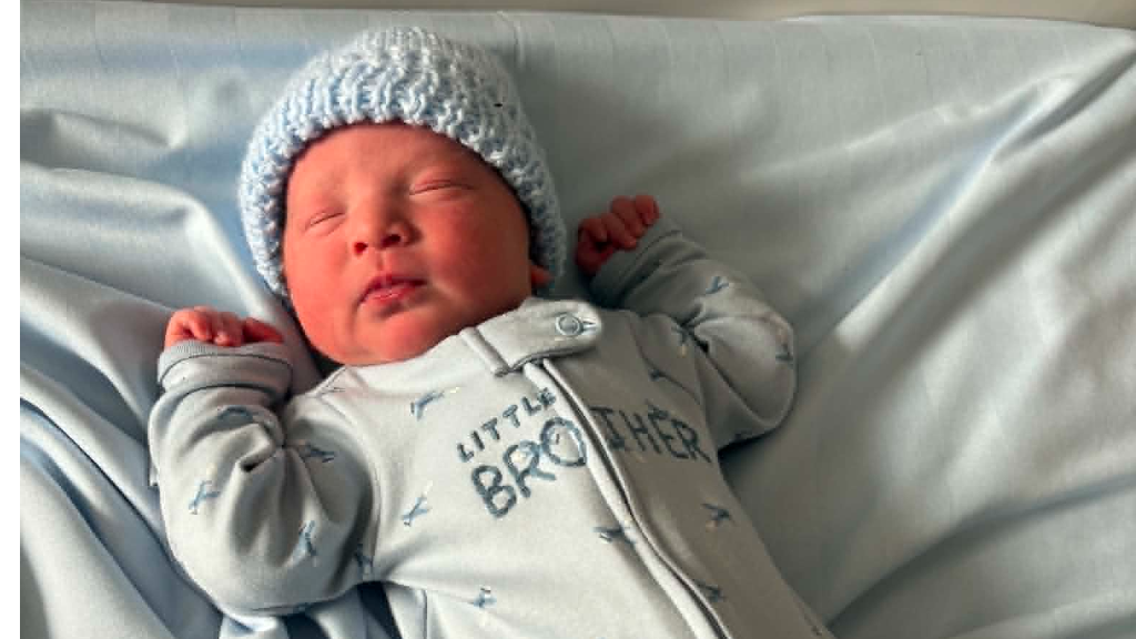 A baby wearing a blue hat and blue onesie that says "little brother" on the front.
