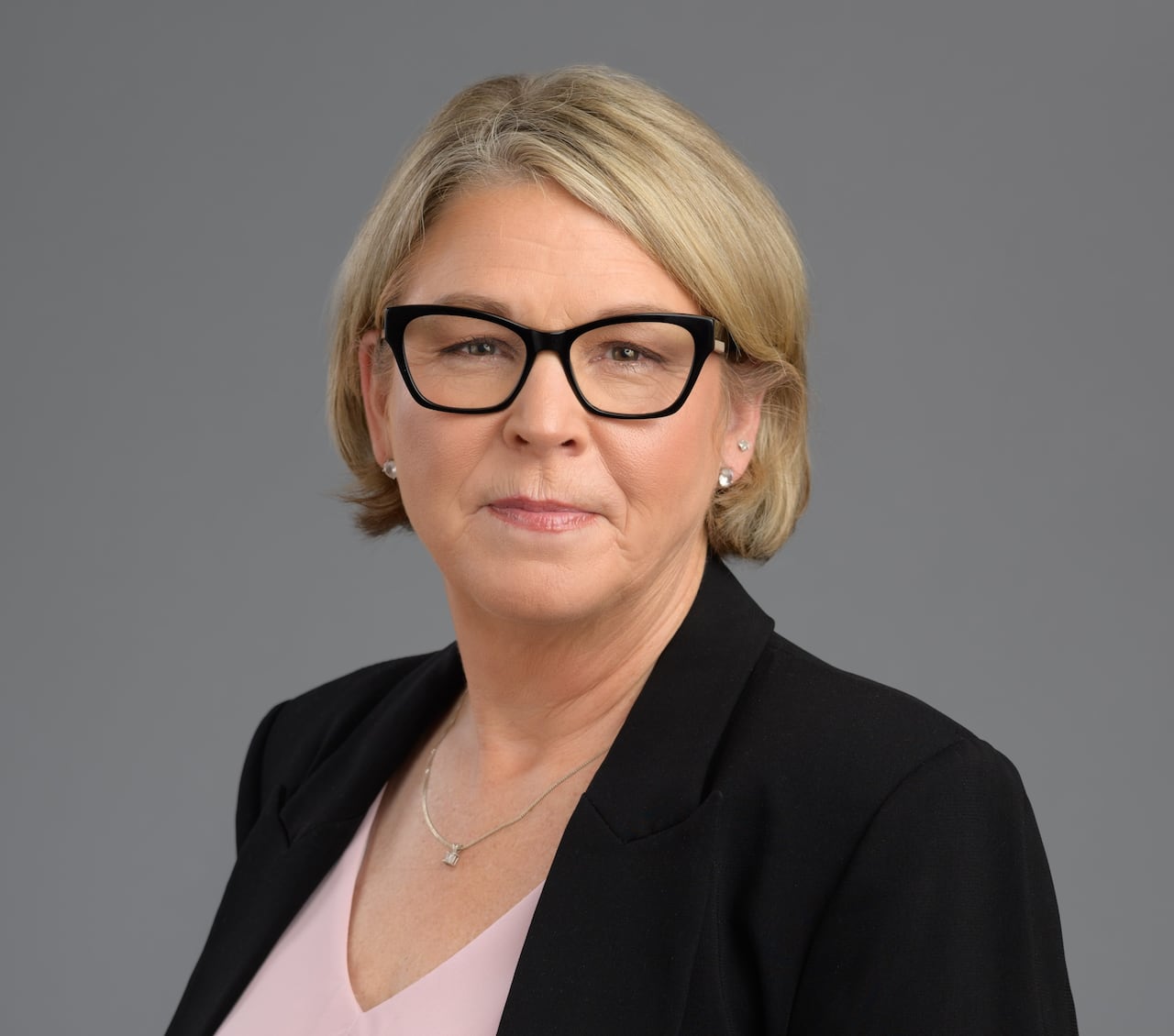 A blond woman in a pink shirt wearing a black blazer and black glasses.