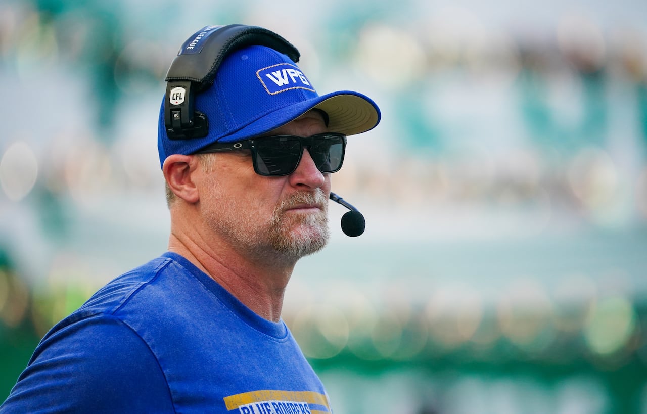 A football coach is seen from the shoulders up, wearing a blue ball cap and a headset.