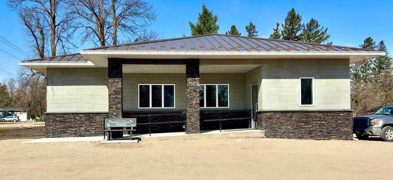 A brown and tan building with windows and pillars on the front has a bench at the entrance. The gorund is covered in gravel.
