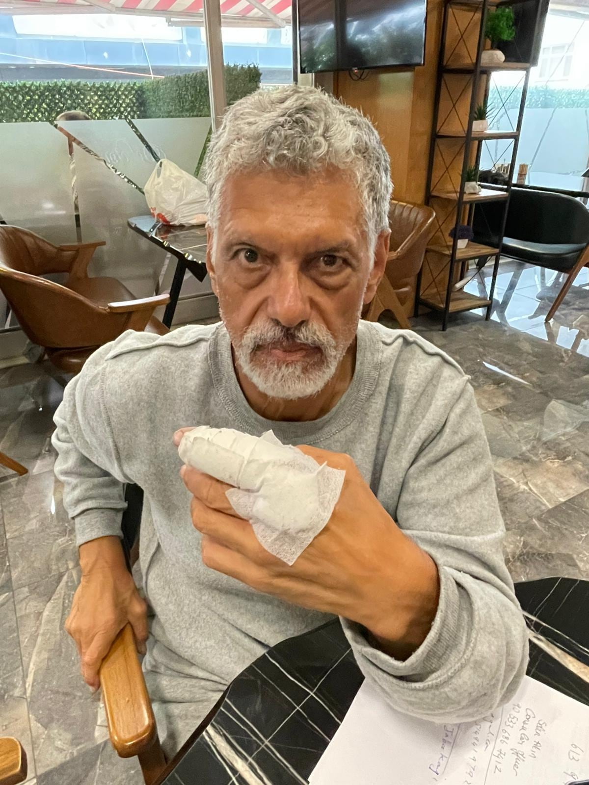 A man sitting down, wearing a grey jumpsuit, holds his hand up that is wrapped with gauze.