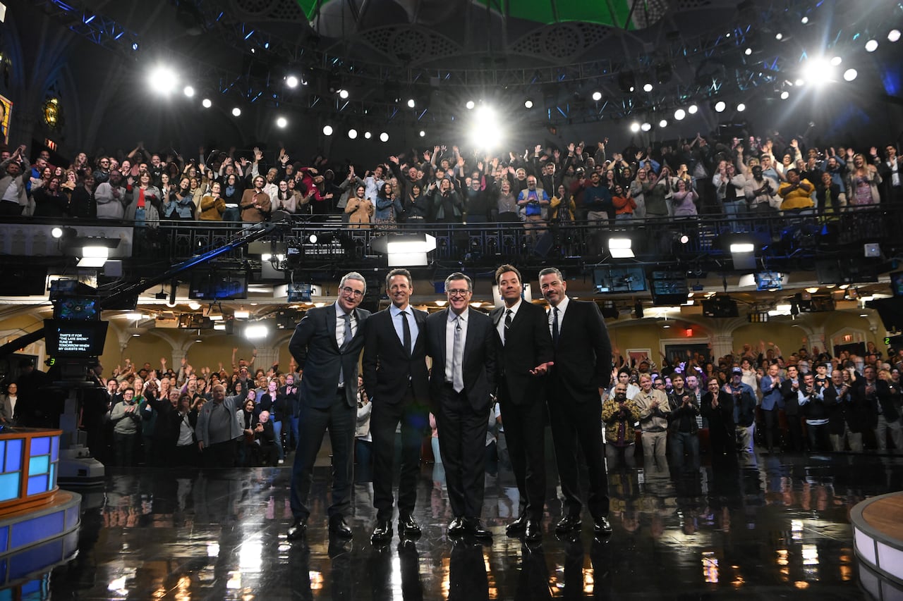 Five men in suits stand together in front of a big audience.