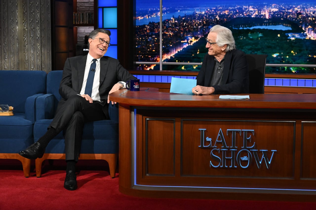 Two men in suits facing one another. One on chair and one behind desk.