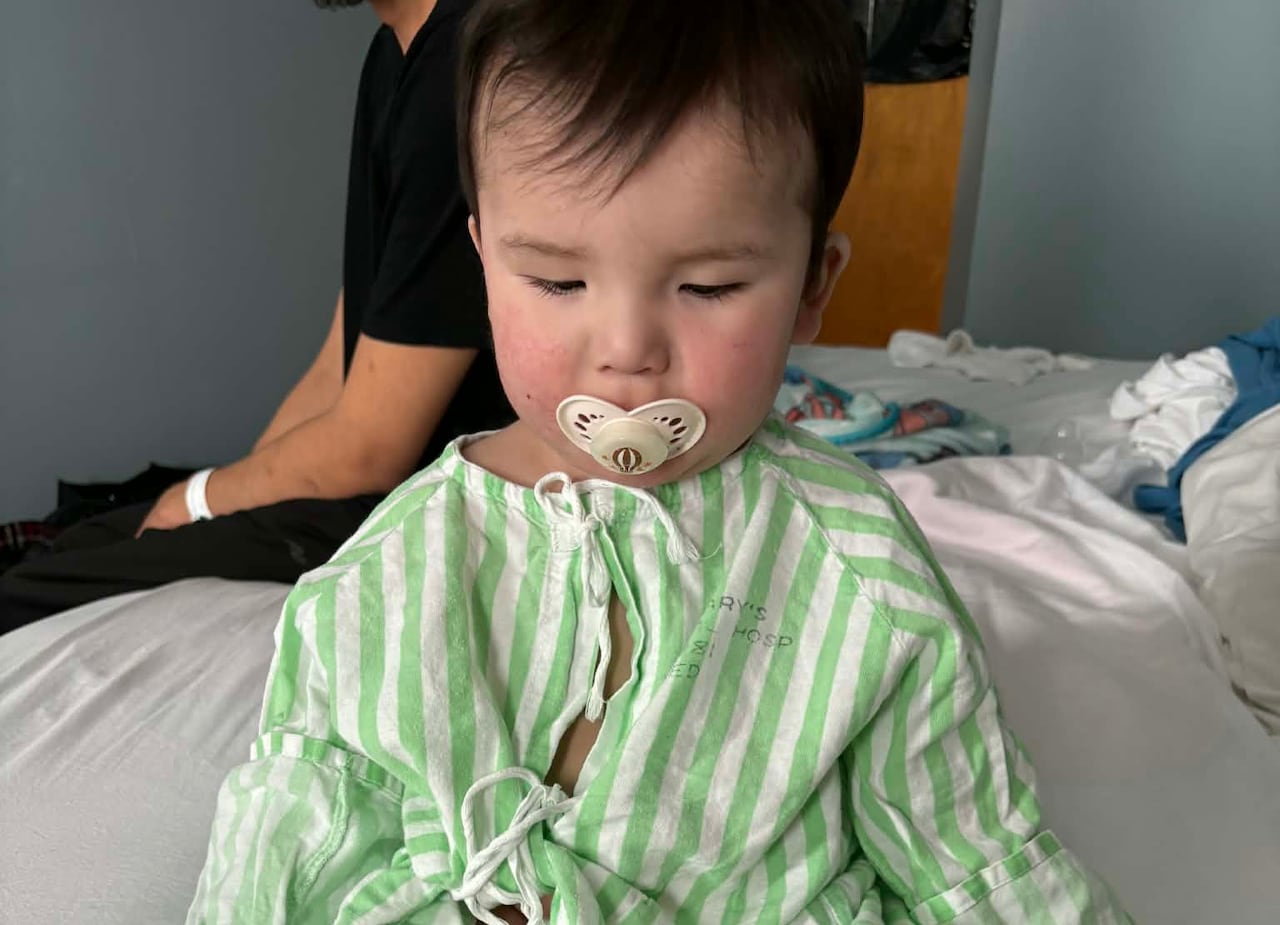 A chubby baby wearing a green-striped hospital gown seated on a bed