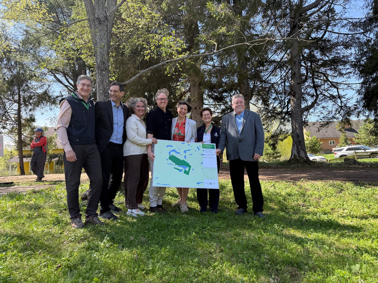 Seven people stand outside under the shade of a tree holding a big map on a board showing the protected area around Lac Tamaracouta.