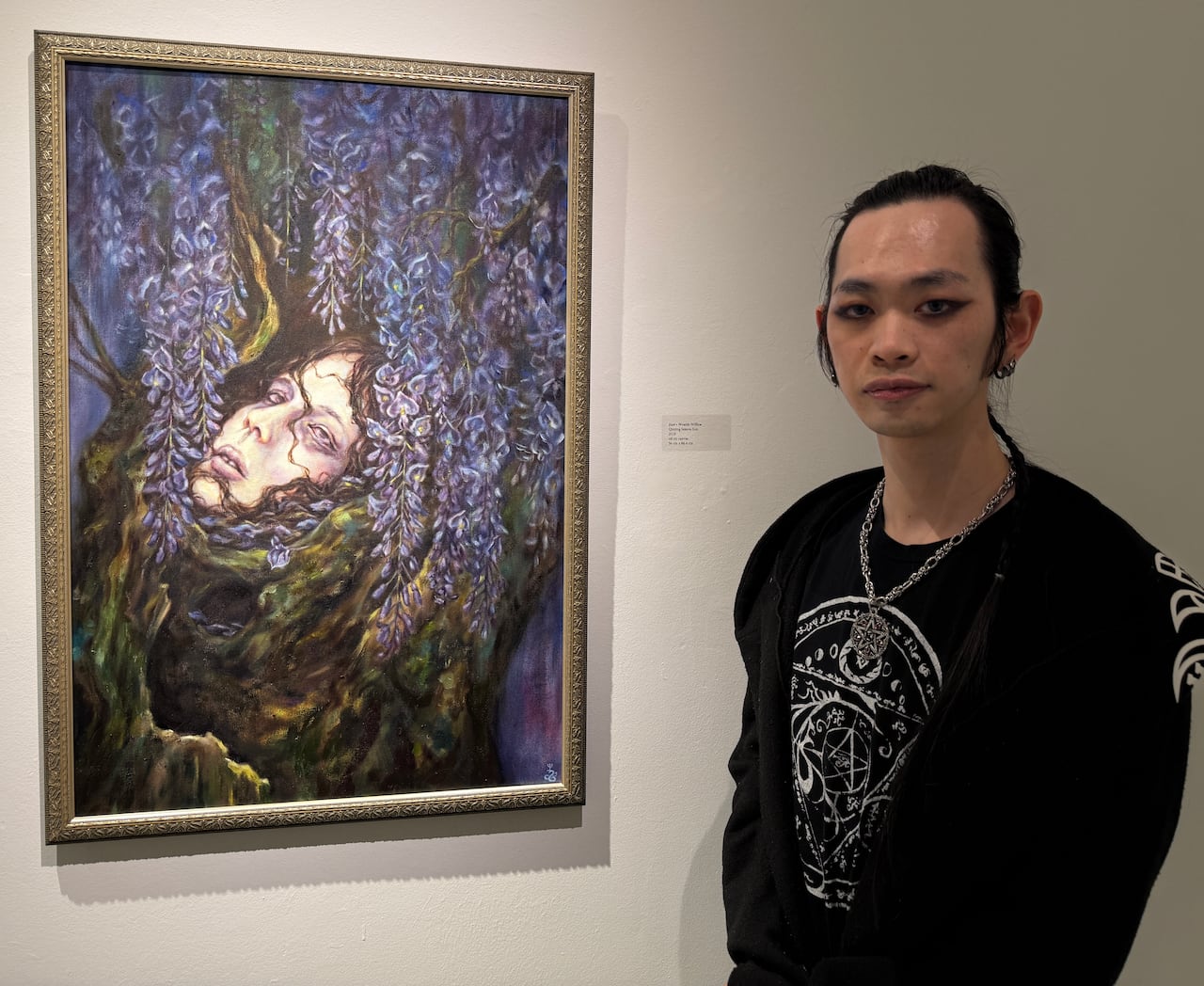 A man in dark clothing stands next to a framed painting on a gallery wall.