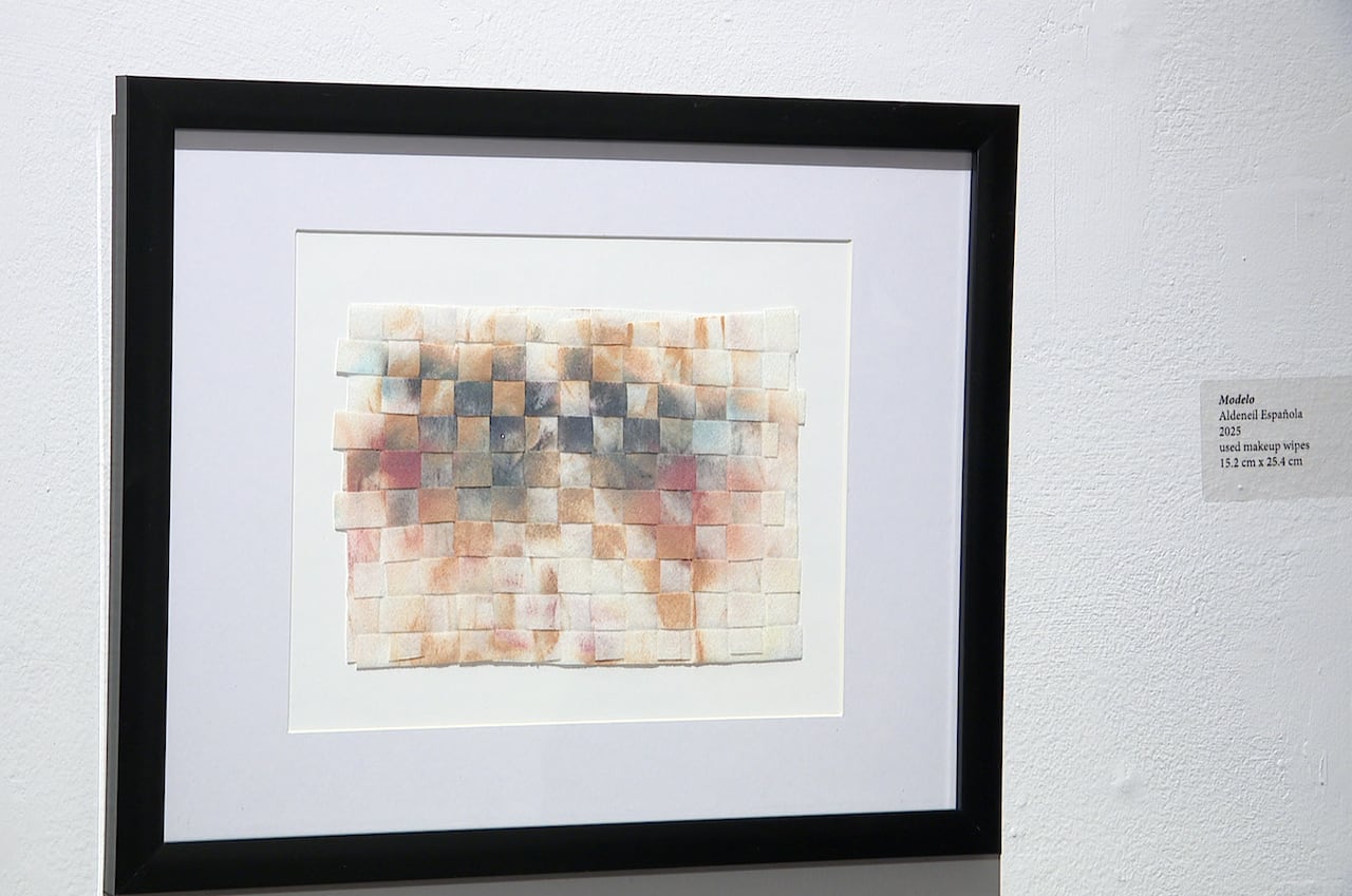 A framed artwork made of smudged and stained squares hangs on a gallery wall.