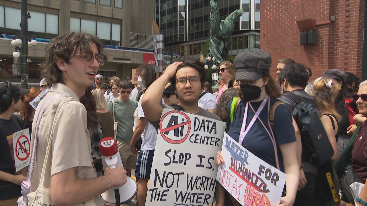 A crowd of people stand outside holding protest signs, including one that says “AI data center slop is not worth the water.”