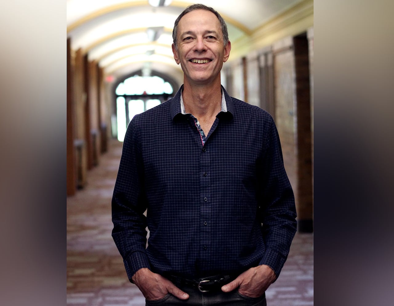 A man stands with his hands in his front pockets, smiling and wearing a dark, collared shirt.