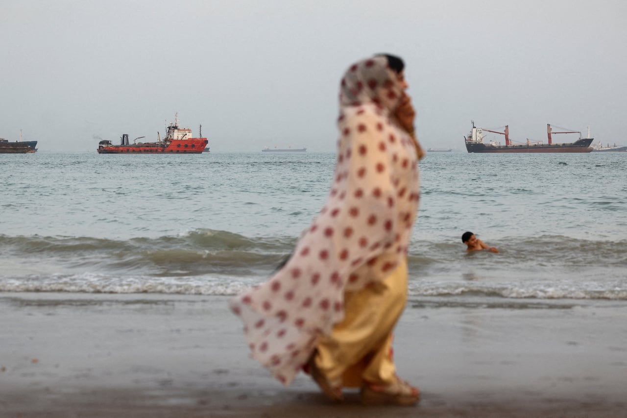 A woman wearing a shawl walks on a beach with large boats anchored in the distance.