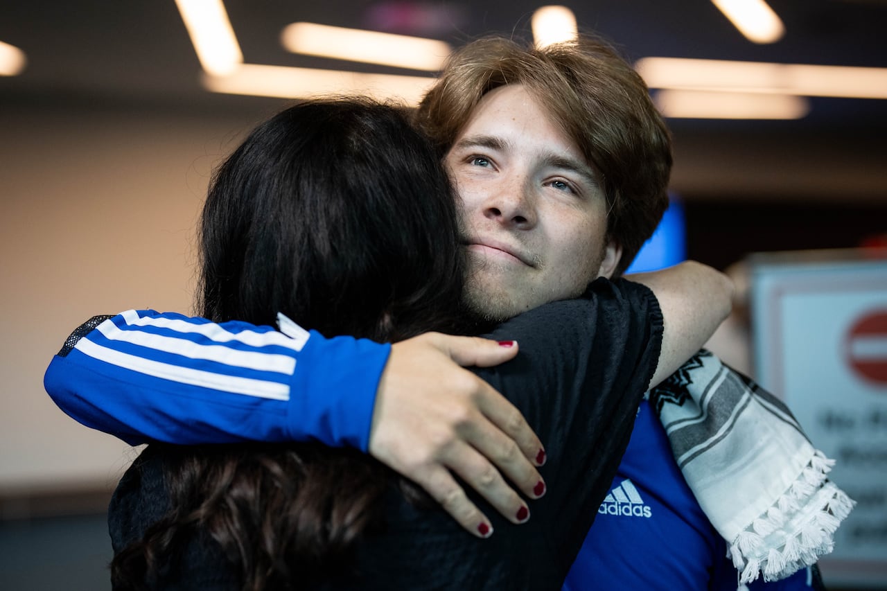 A young man in a blue shirt hugs another person inside an airport arrival area