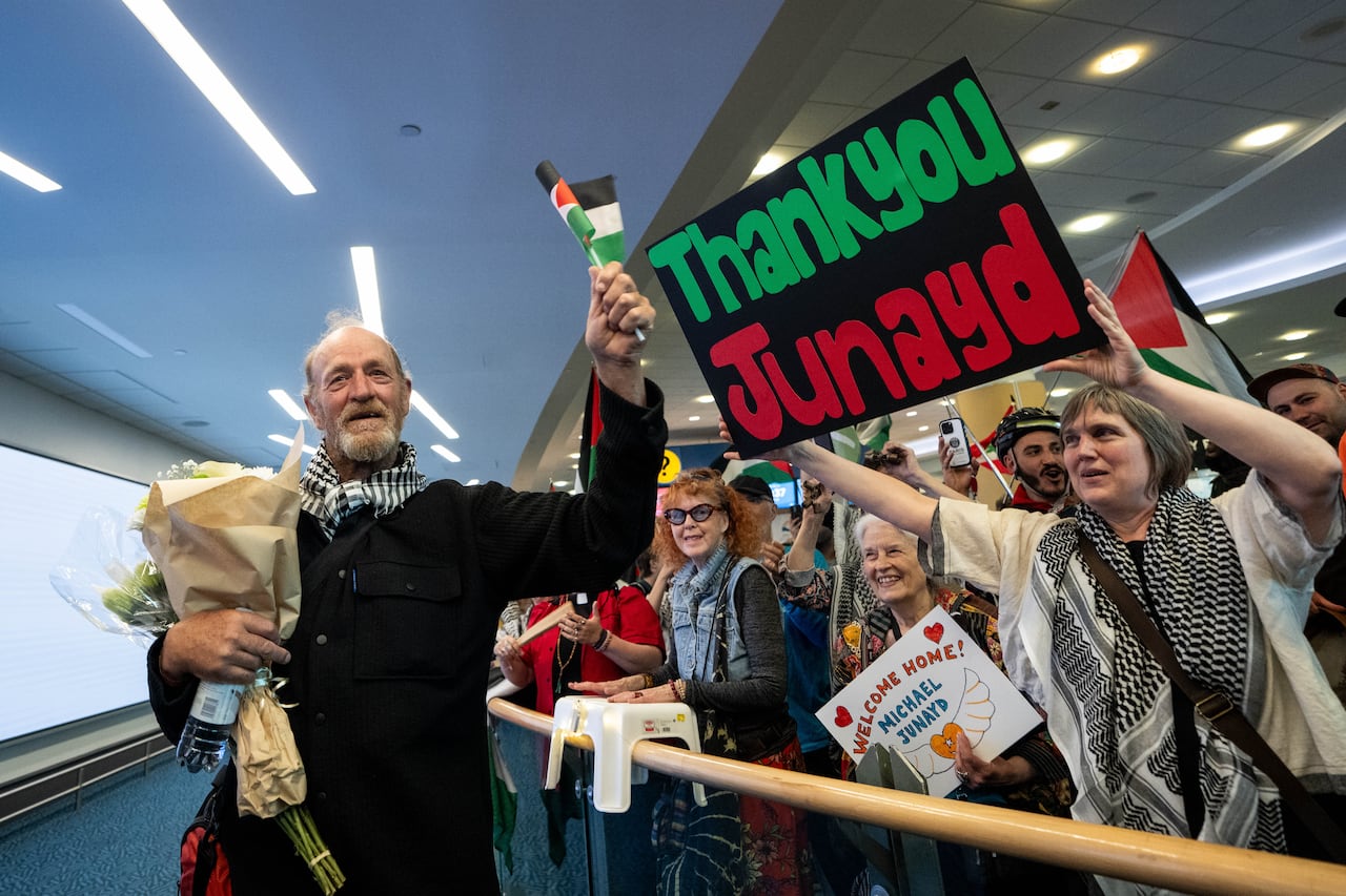 A man holding flowers waves a small Palestinian flag while supporters hold signs and Palestinian flags inside an airport arrival area.