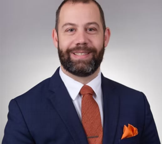 A bearded man in a suit and tie looks at the camera.