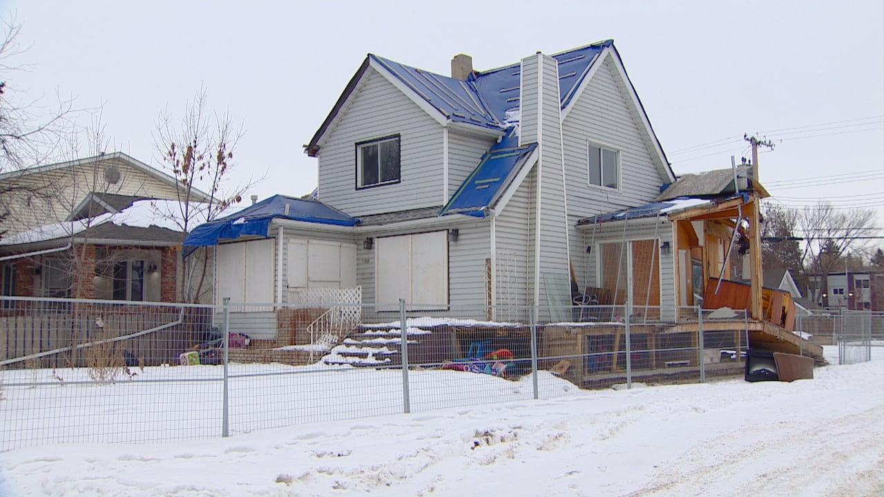 A blue house without roof tiles, and with boarded-up windows and with parts of it without siding is surrounded by a fence with snow around it.