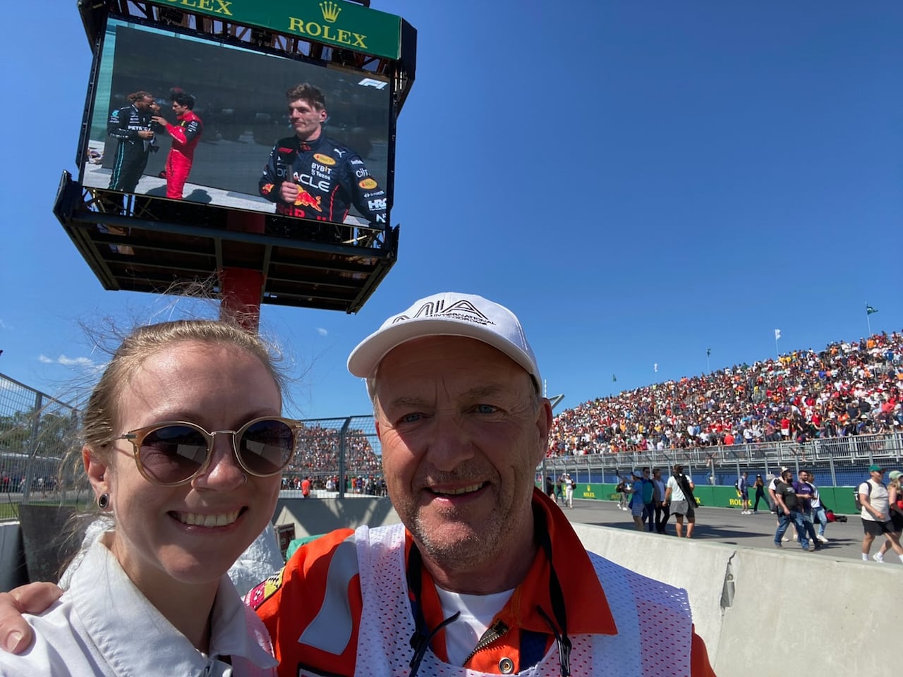 An older white man with a younger white women in front of filled stands at a race track