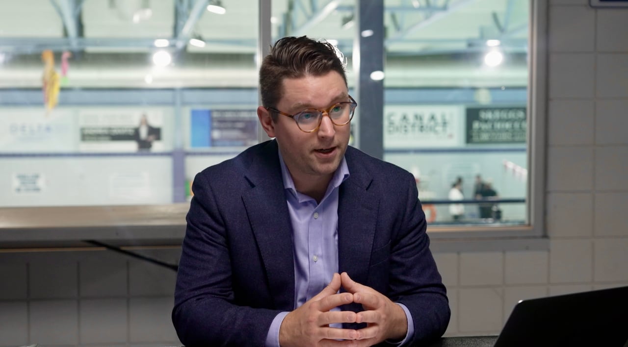 Man in suit with glasses speaks while sitting at a table.