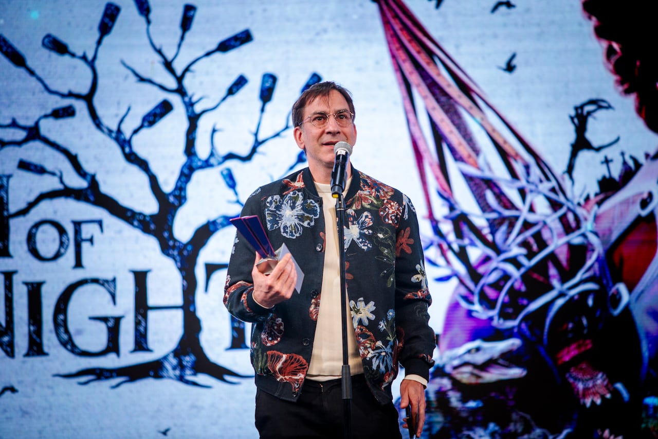 A man in a flowery jacket speaks into a microphone on stage, while holding a trophy.