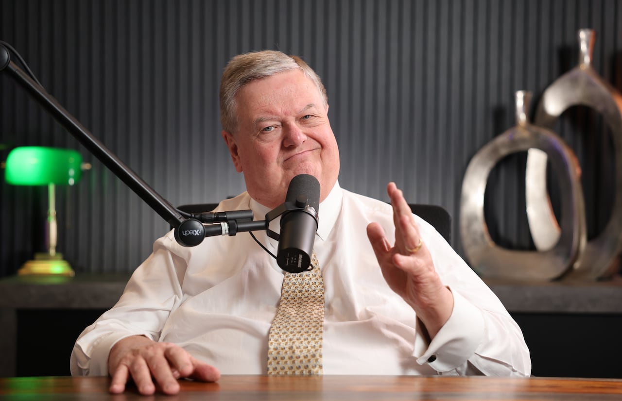 A man in a dress shirt and a tie sits at a desk behind a broadcast microphone.