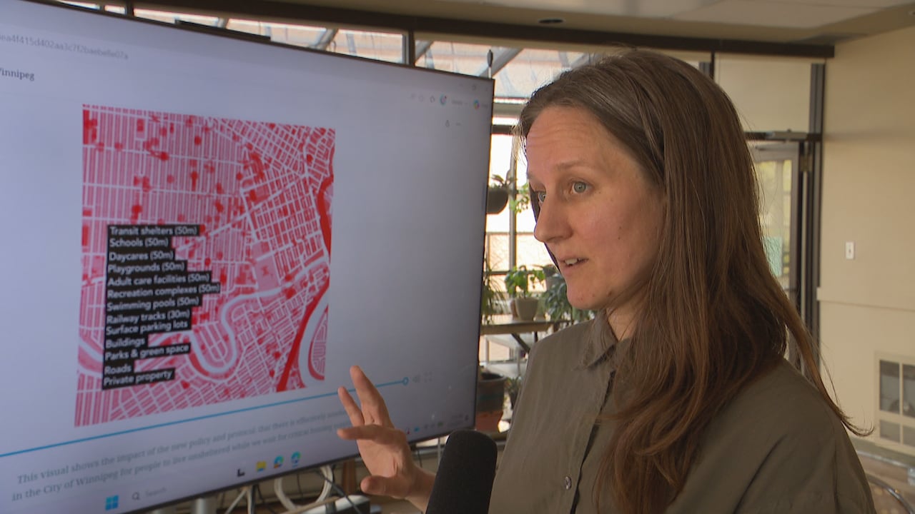 A person with long hair, wearing a green-ish collared shirt, gestures with their hand while speaking in front of a red map.