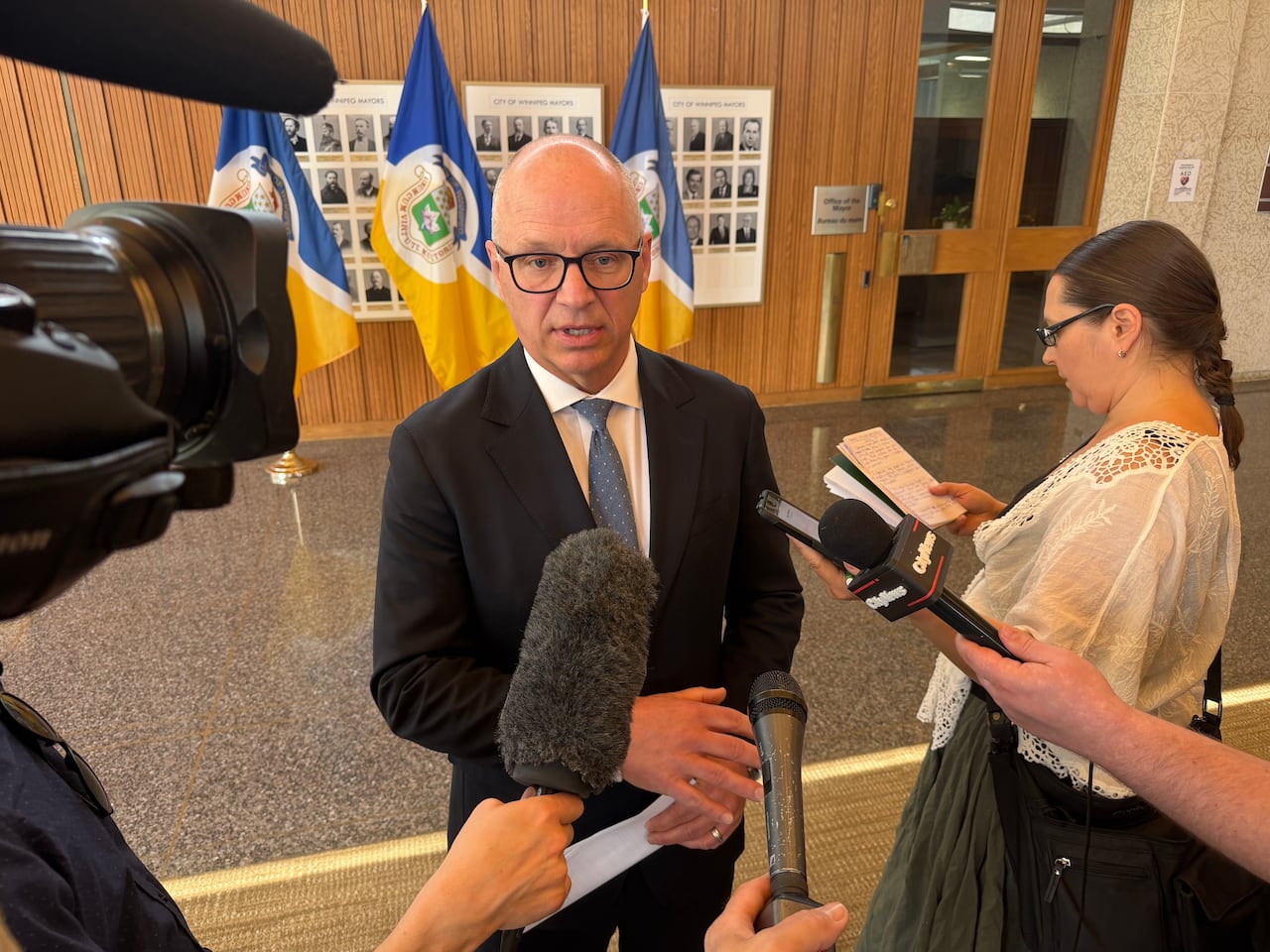 A man in a blue tie and dark suit speaks with reporters at a city hall.