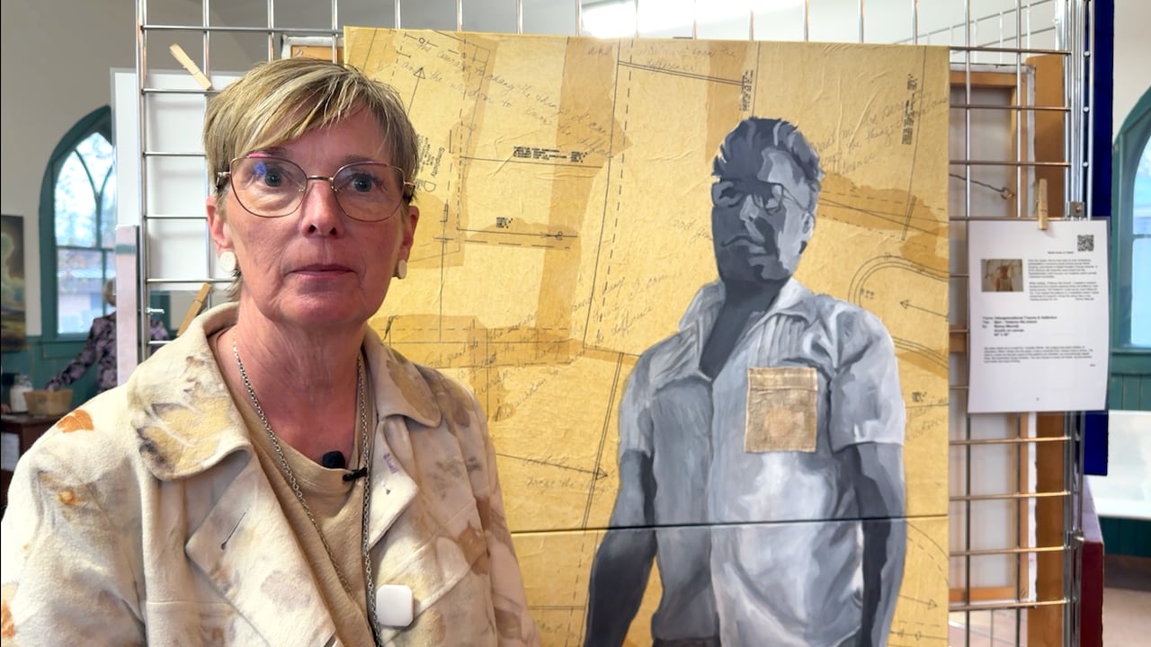 A short-hair blonde woman stands in front of a painting with a greyscale man on a background of aged and yellowed sewing patterns.