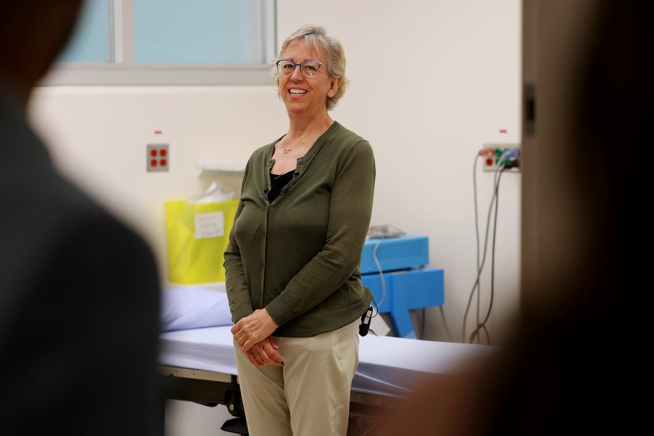A woman stands and smiles while standing in front of a hospital bed.
