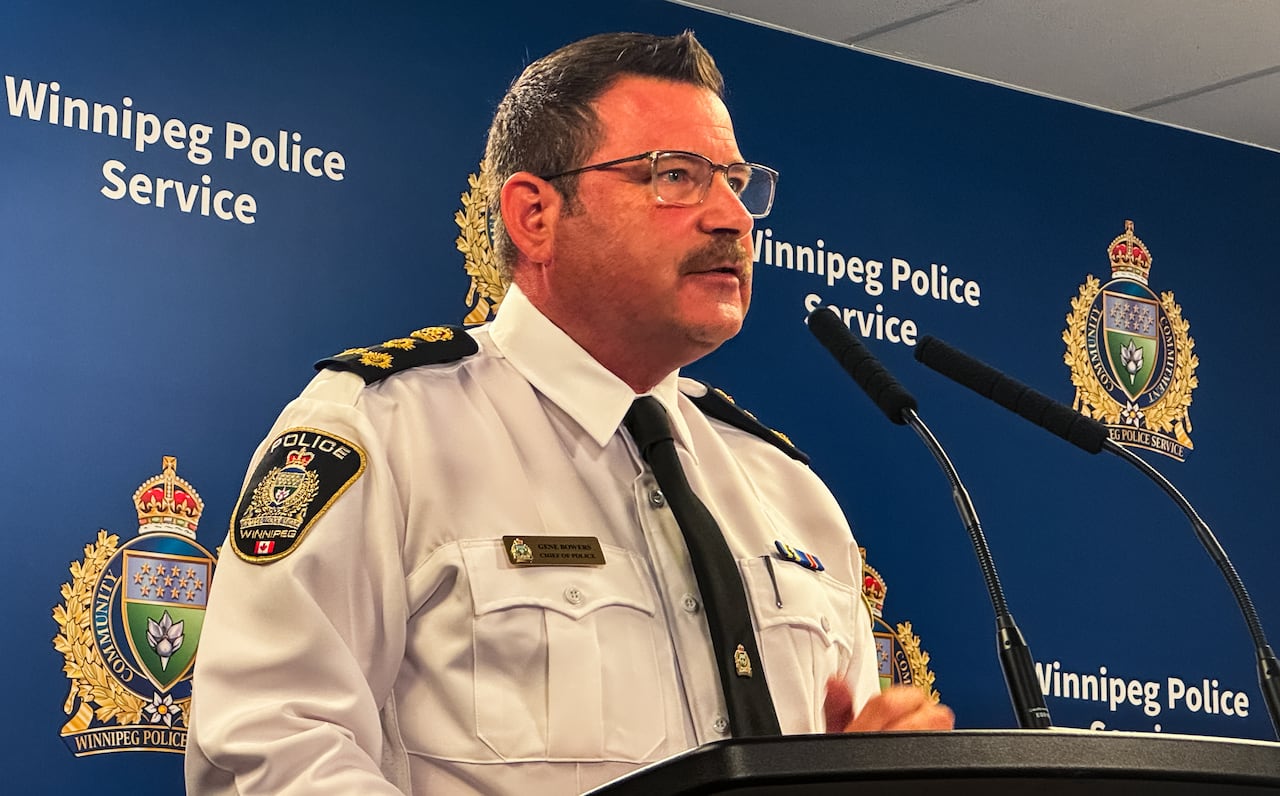 Man with a moustache wears a white police uniform shirt and stands behind a podium speaking into a microphone. A banner behind him reads "Winnipeg Police Service."