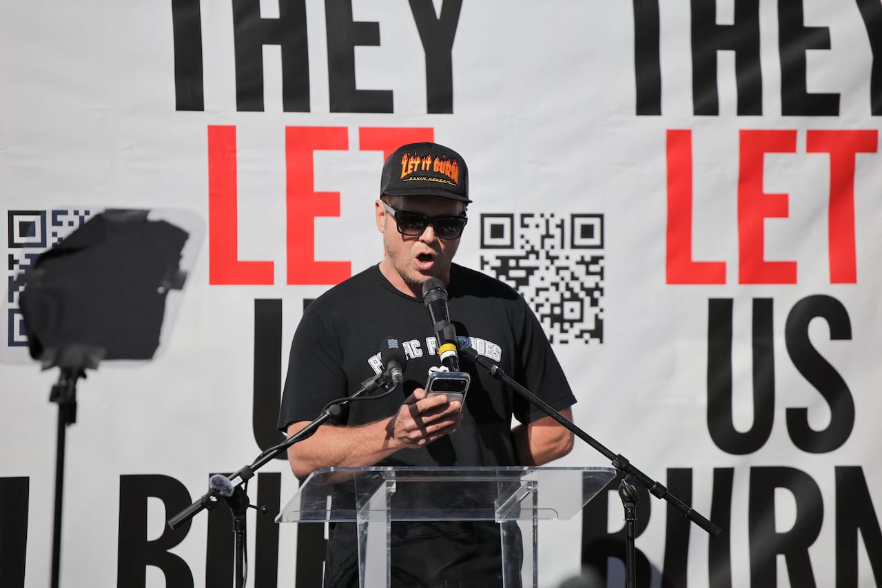 Man in hat reads off a phone while standing in front of a banner with a QR code and the text THEY LET US BURN.