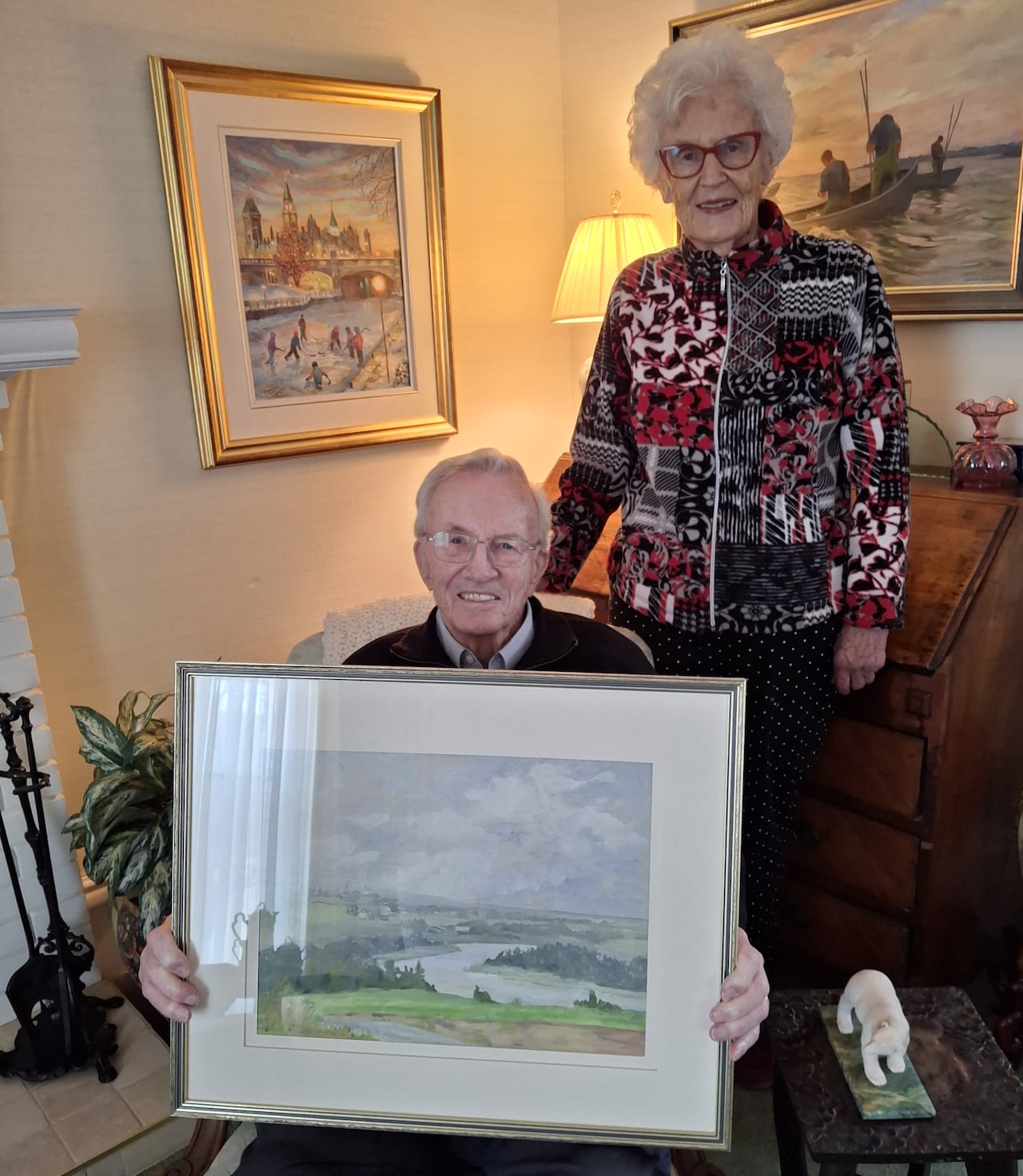 An elderly man seated and an elderly woman standing hold a framed landscape painting in a living room.