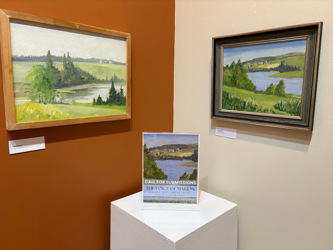 A close-up corner view of an art gallery featuring two framed landscape paintings hanging on contrasting orange and beige walls, with a call-for-submissions flyer on a white pedestal below them