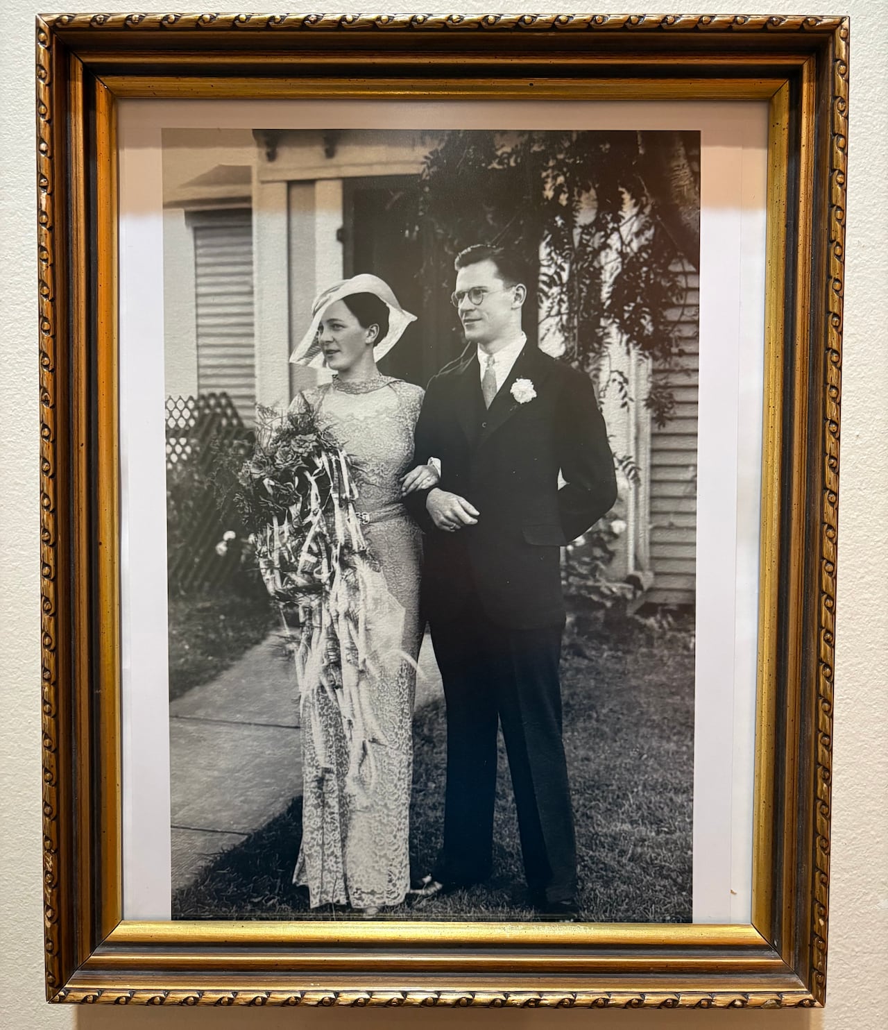 A framed, black-and-white vintage photograph of a bride in a lace gown and wide-brimmed hat holding a large bouquet next to a groom in a suit and glasses.