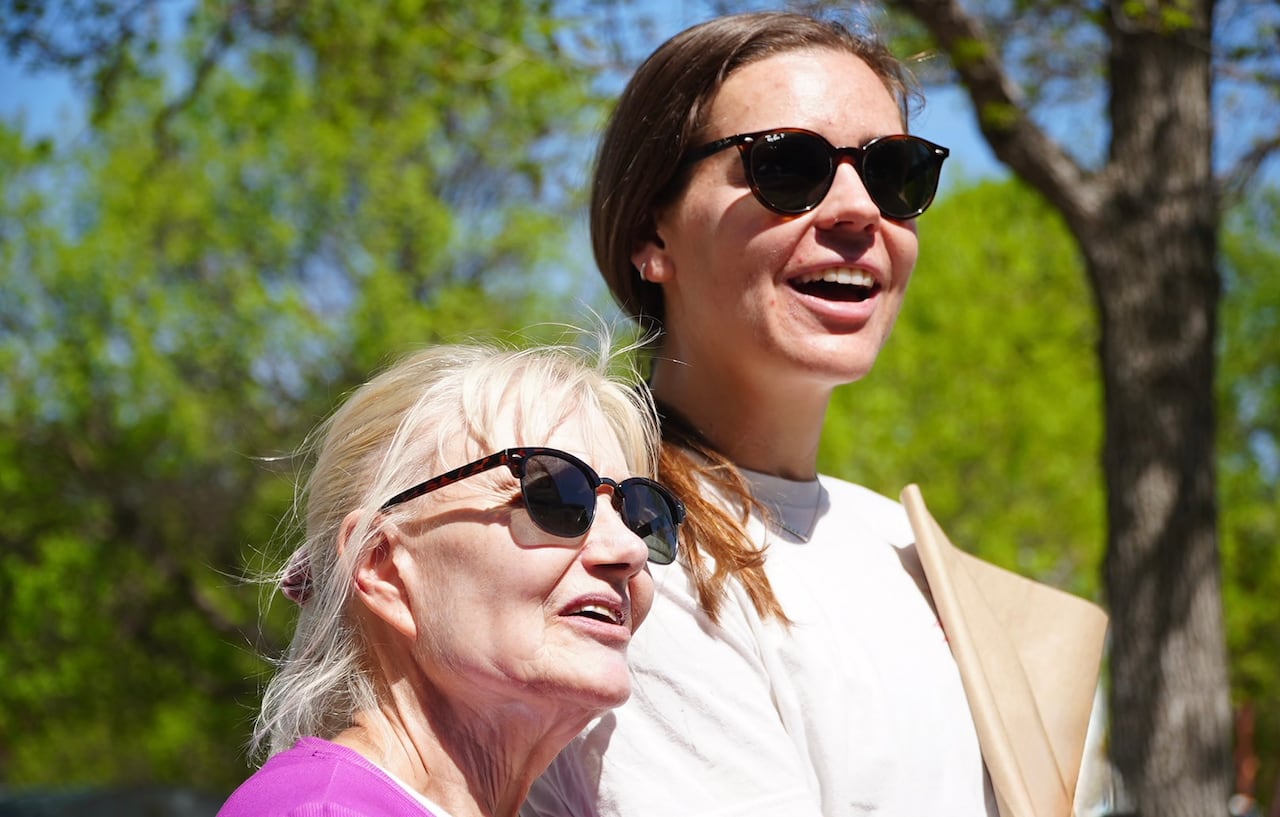 An older woman with white hair stands next to a younger woman with brown hair. Both wear sunglasses and smile
