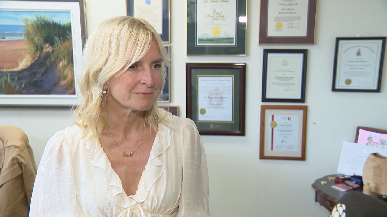 Blonde woman in white blouse stands in front of wall with certificates hanging on it