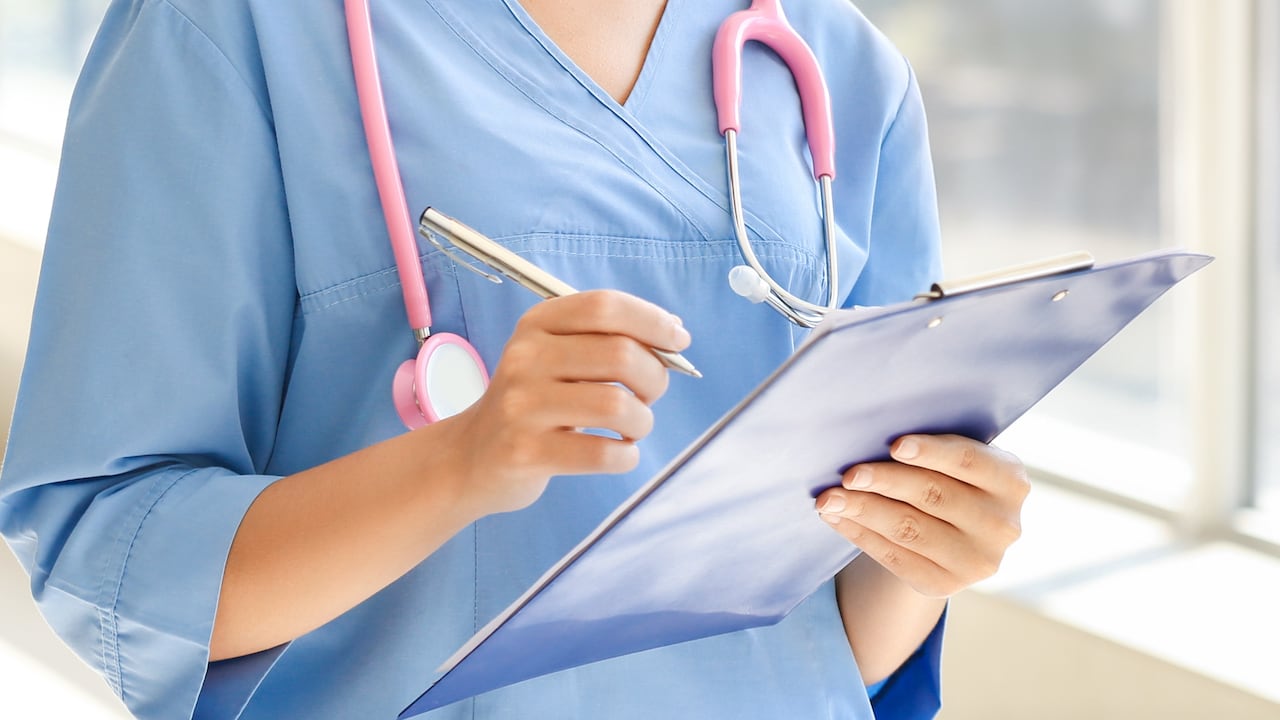 Young African-American nurse wearing blue scrubs, with stethoscope around her neck, holds clipboard in left hand and pen in right hand.
