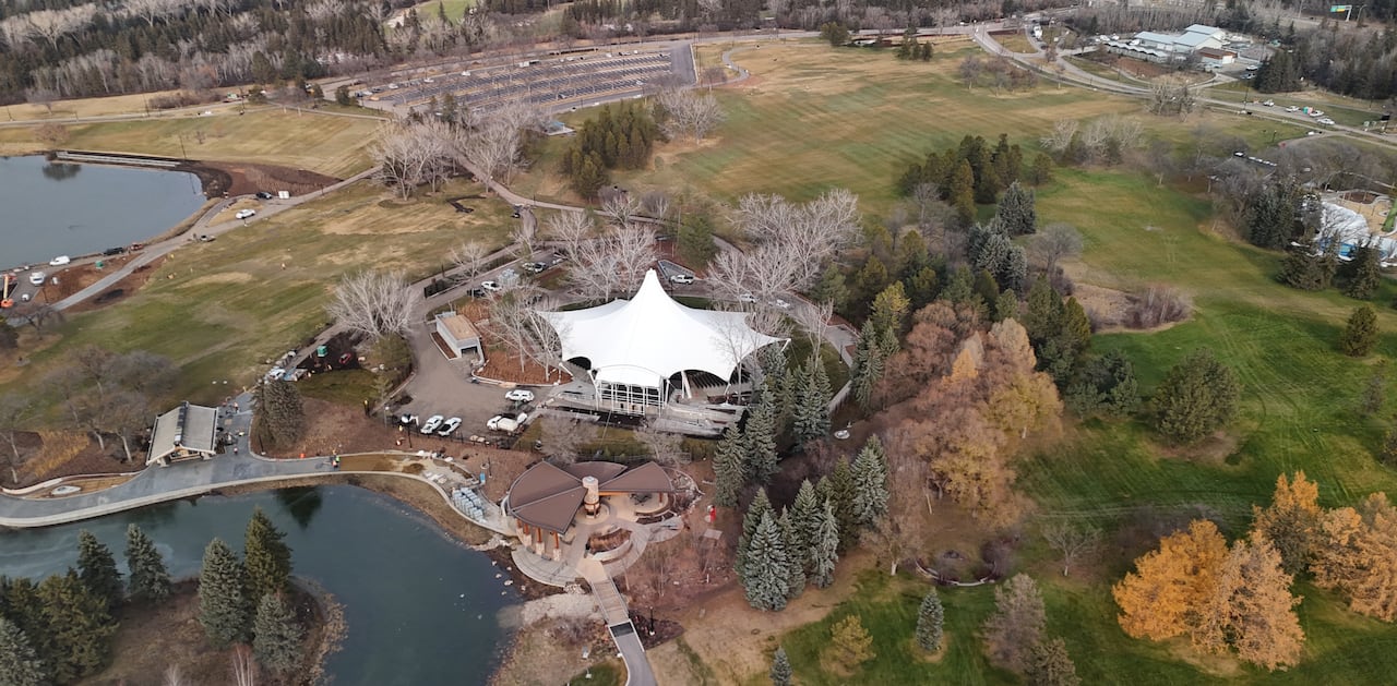 A drone shot of a park. There is a large structure in the centre of the park. Grass fields and a paved parking lot are seen in the background.