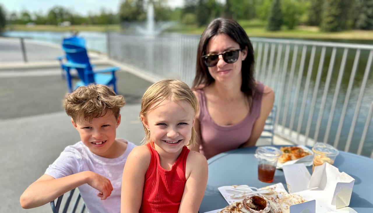 A women and her two children sit at a table by a small lake. A fountain and trees are seen the the far background.