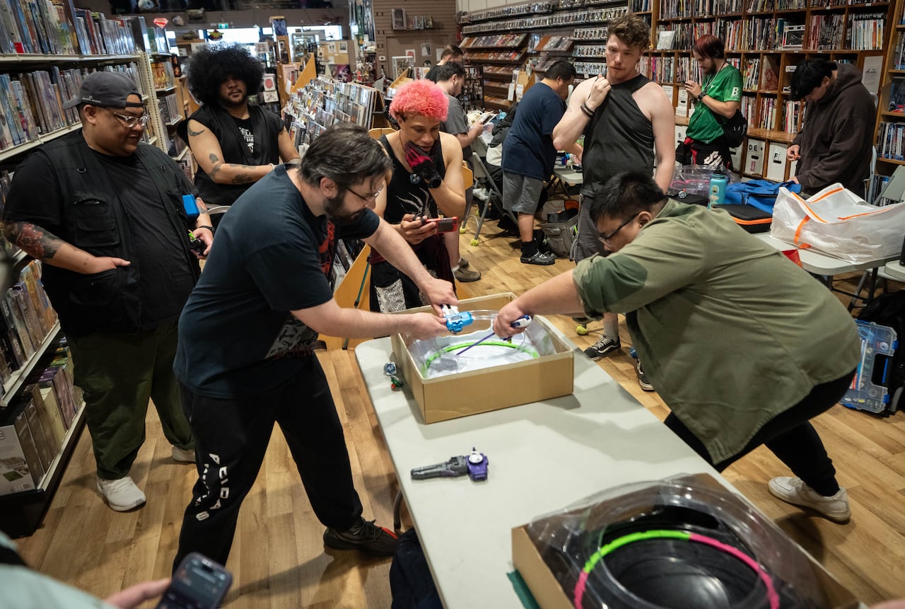 A group of people in a card shop gather around two people spinning tops in an enclosed box.